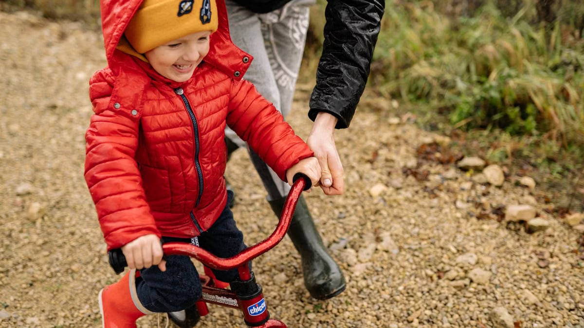 Joyful child riding a bike with parent on a gravel path, enjoying family time outdoors.